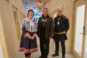 Three people standing in wooden hallway, two with traditional Sami clothes