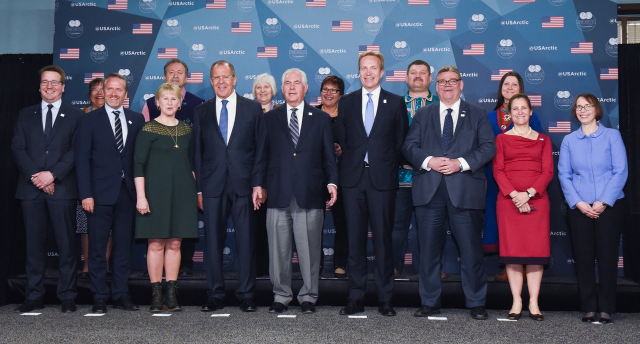 A group of men and women standing in front of a wall with US flags and Arctic Council logos.