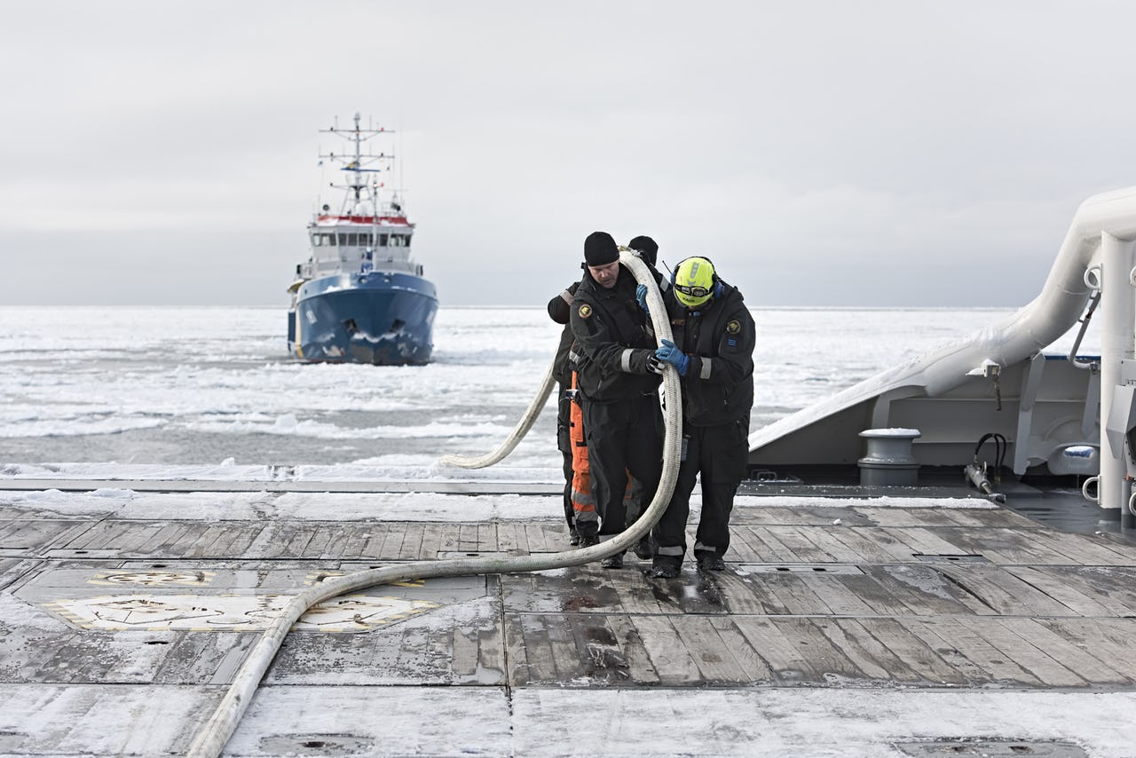 Three men carrying a hose on board a ship with icy waters and another ship in the background.