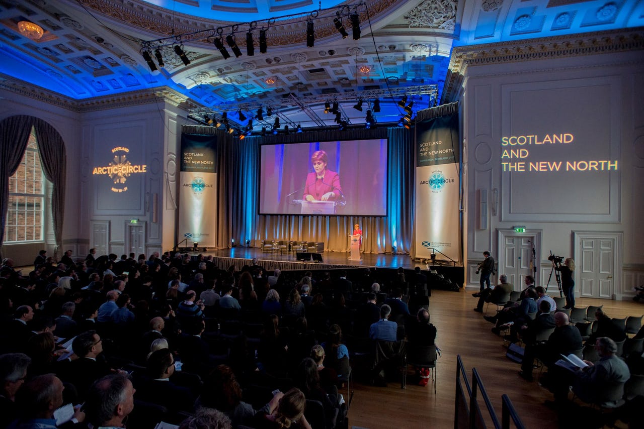 Crowd of people in a large conference hall in Scotland with a woman on a large screen in the background