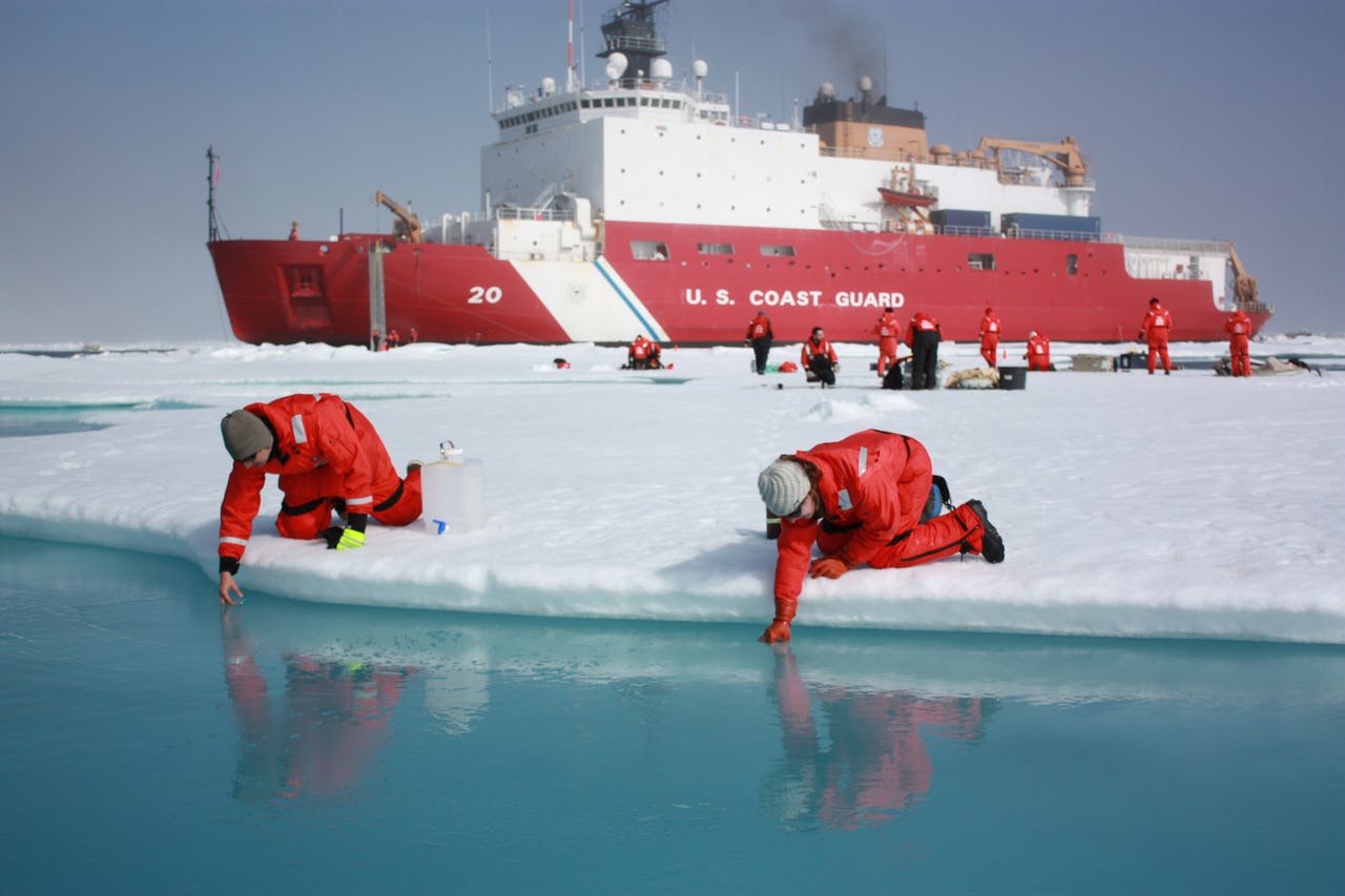 Two men scooping water from melt ponds on sea ice with icebreaker in the background