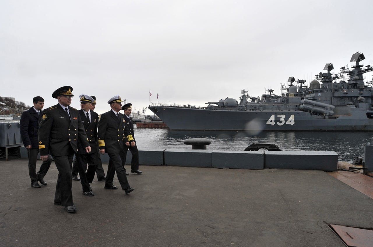 A group of men in uniform walking in a port area with a military ship in the background.