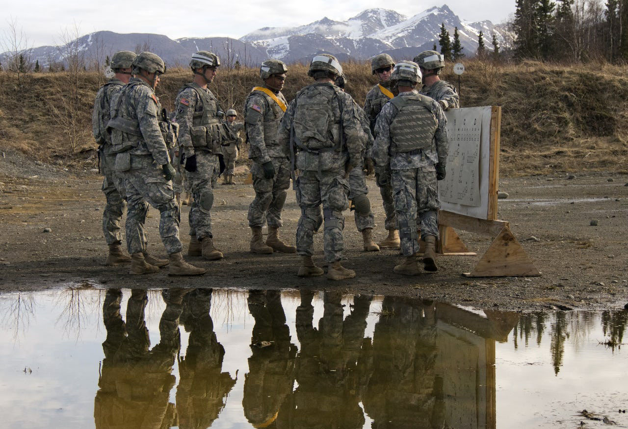 Group of men in uniform standing against a background of snowy mountains. Group reflected in a large puddle.