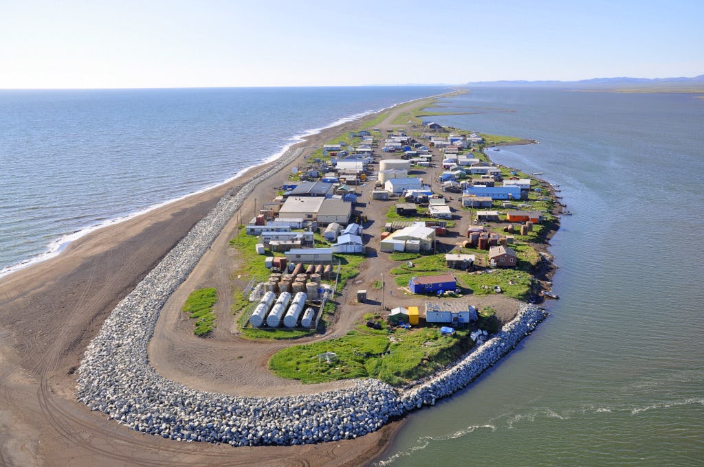 Buildings and rock wall on a dirt spit with grass surrounded by water.