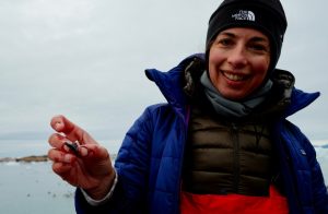 Smiling woman in blue jacket and black hat holding a small piece of whale in her hand