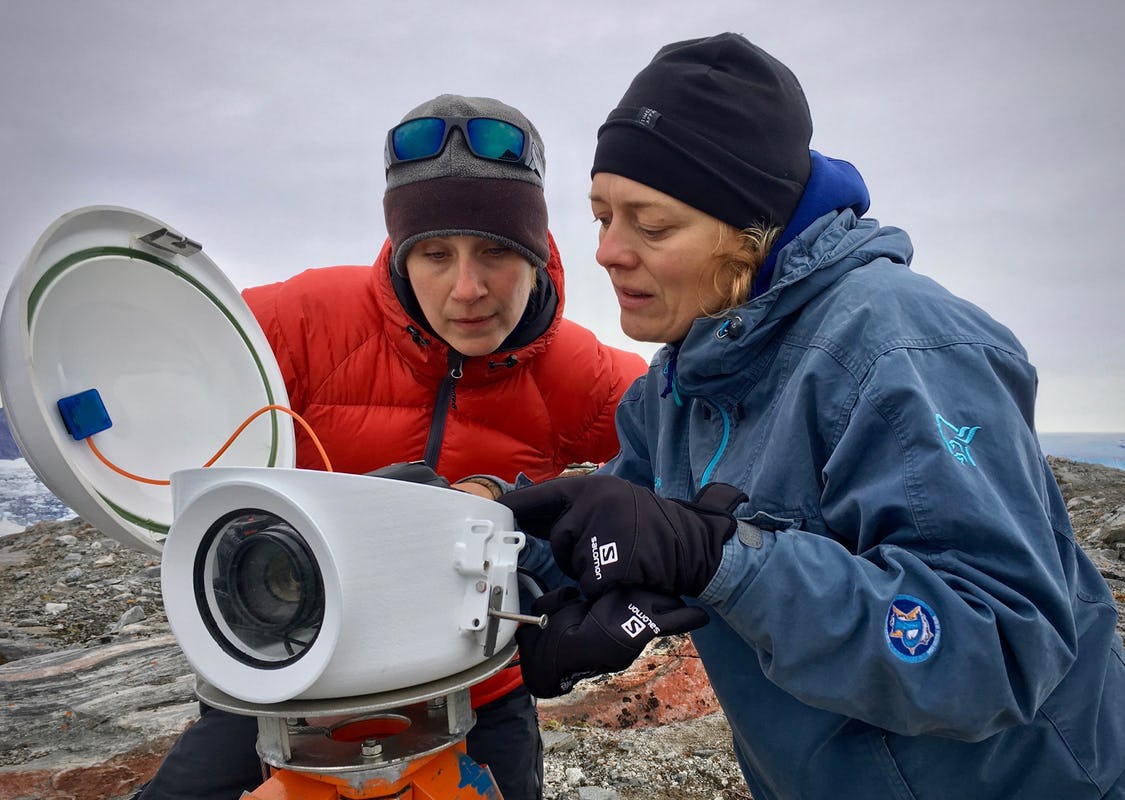 Two women in red and blue jackets with woolly hats examine a big grey camera