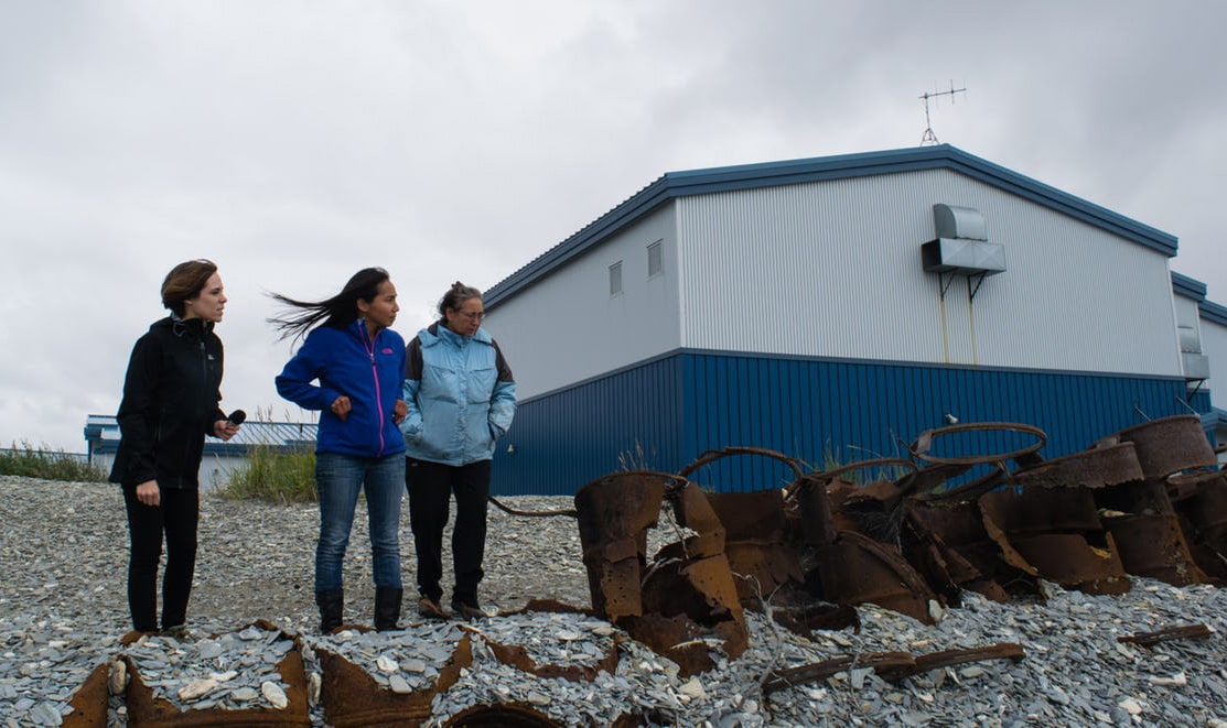Three women standing on a stony beach with blue-grey building in the background