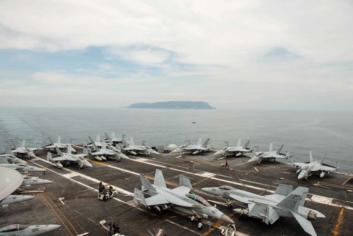 Nuclear-powered supercarrier with fighter jets on deck with island in the background