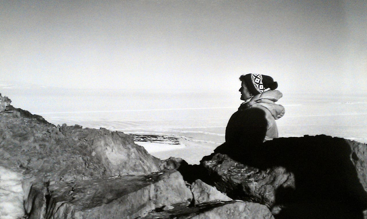 A woman in a cold-weather coat and hat sits on a rock and looks out over an icy landscape in Antarctica