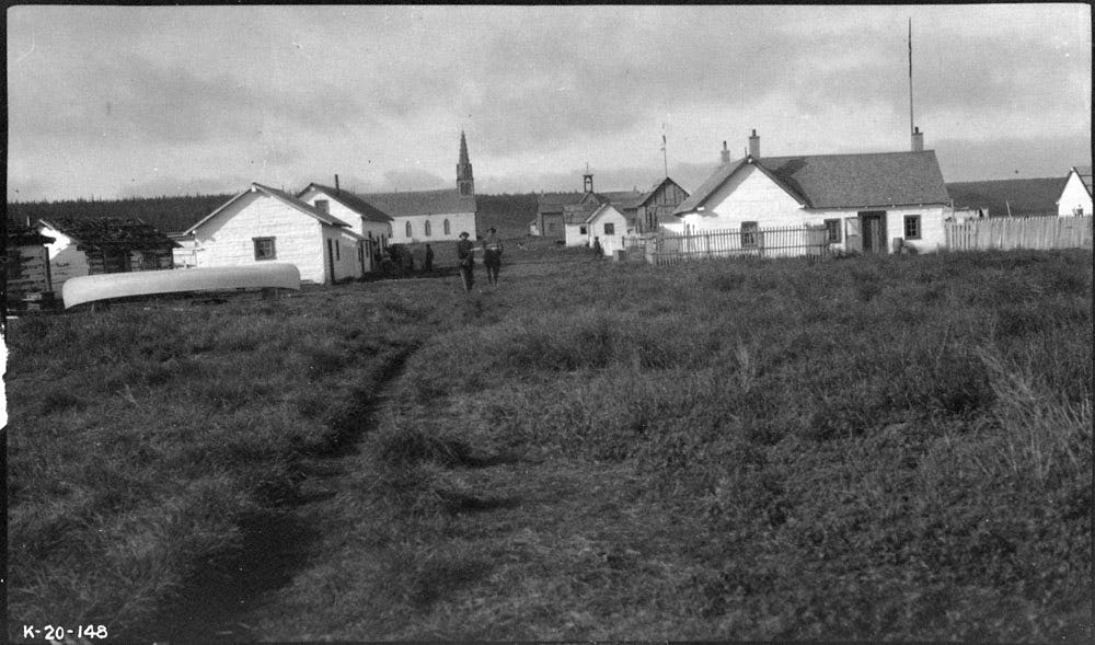 Black and white photograph of Fort Good Hope in the Northwest Territories, Canada, showing a small village with a few white houses and a church tower surrounded by grassy land