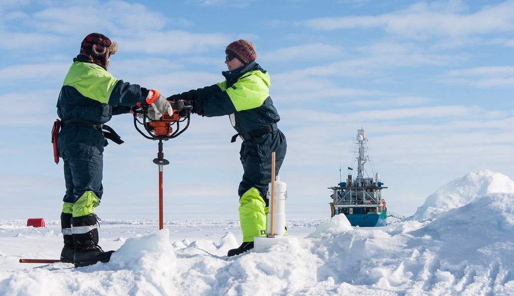 Two women drilling into sea ice in the Arctic. They are surrounded by ice and snow and a research vessel is in the background.