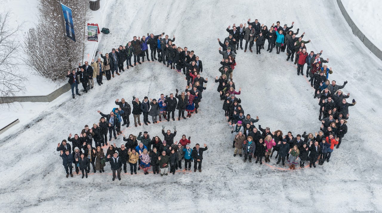 A large group of Arctic Council delegates is in the middle of the photo, standing in the shape of the number “20” on pavement that is white with snow. There are bushes in the upper left hand corner that have grey, bare branches. The people are wearing winter coats and waving up at the camera.