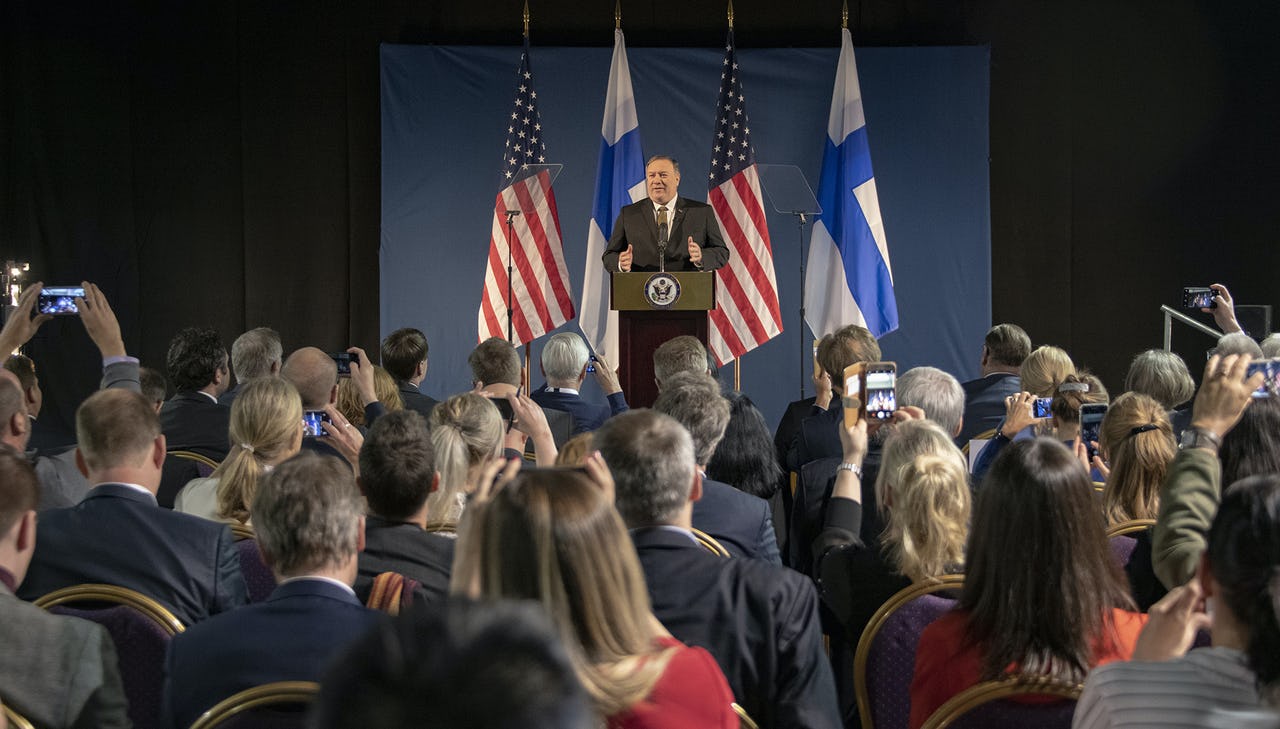 A man speaks at a podium in front of an audience with flags behind him