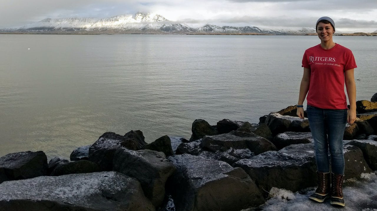 Woman with red shirt and blue trousers standing in front of the sea with mountains in the background