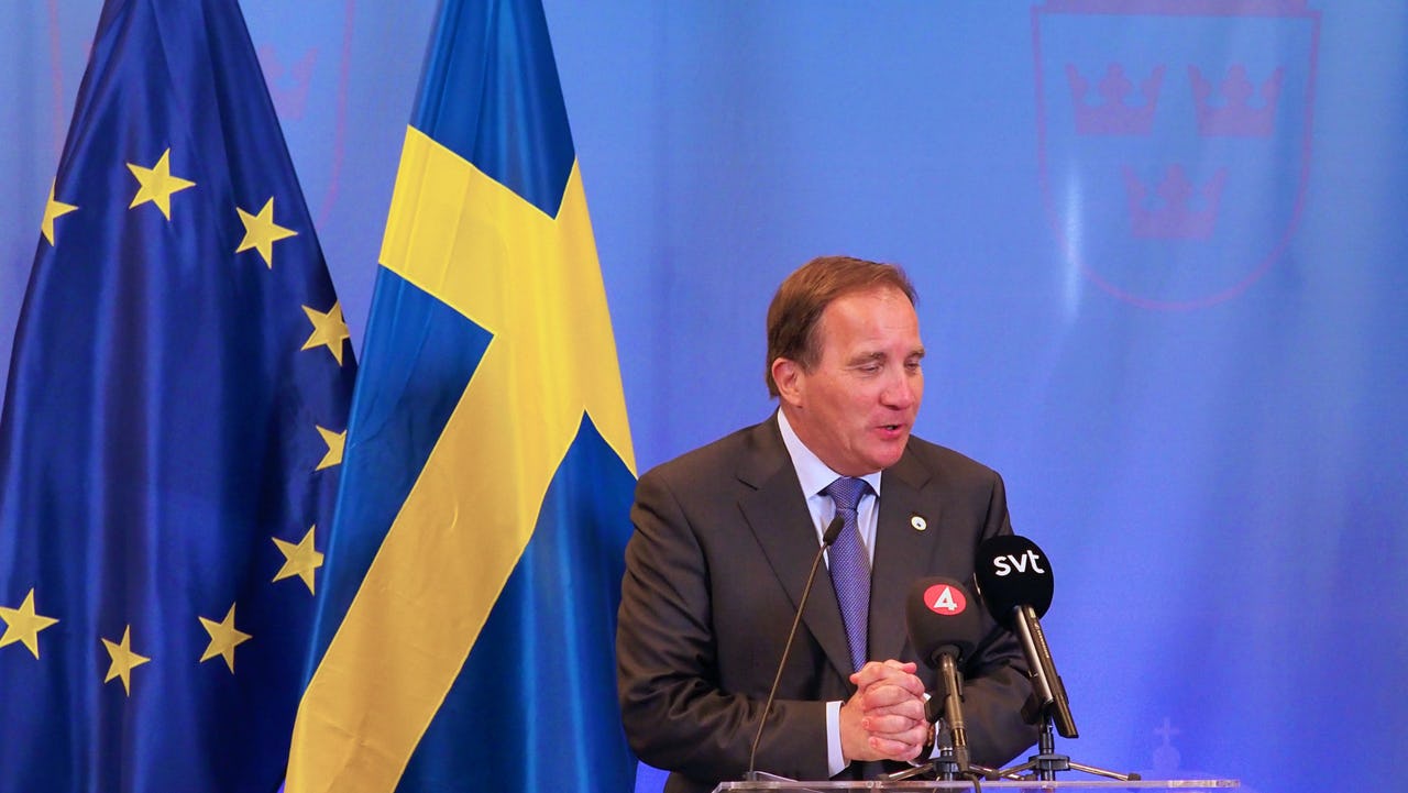 A man standing in front of two flags, both in blue and yellow, and blue background.