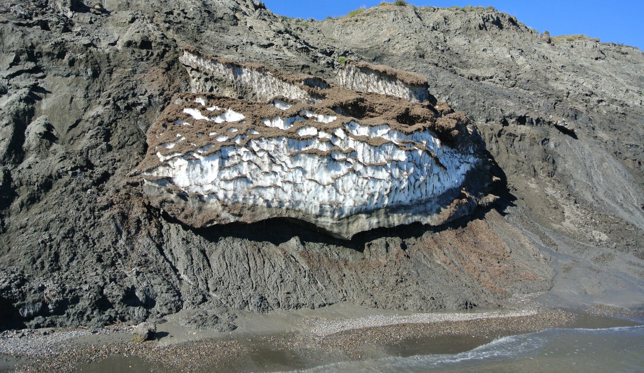 A wall of grey dirt with white ice in the middle and water in the foreground