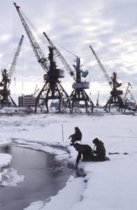 Three Russian fishers fishing on a cloudy day in icy frozen water with industrial plants in background in Krasnoyarsk Krai, Russia, Siberia