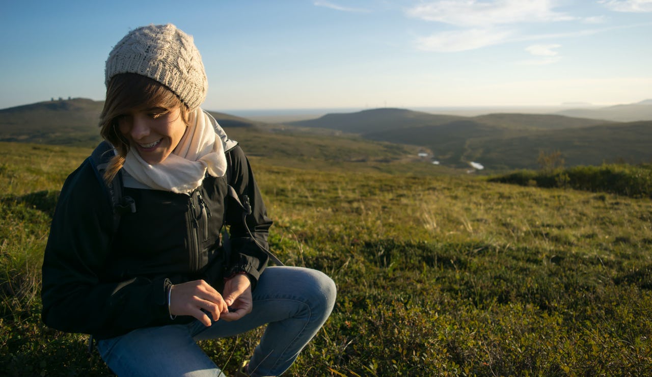 Smiling woman with white beanie, white scarf, and black jacket crouched on hilly grass terrain and hills in the background