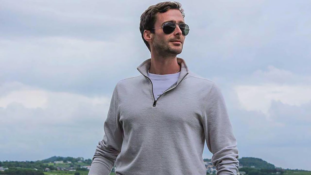 Man with light grey mid-season fleece, dark grey trousers and aviator sunglasses with Arctic sea and green hilly coastline in the background
