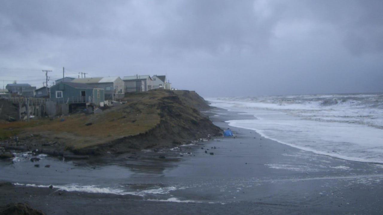 Houses sit on a deteriorating bank of earth near the ocean as a storm rolls in