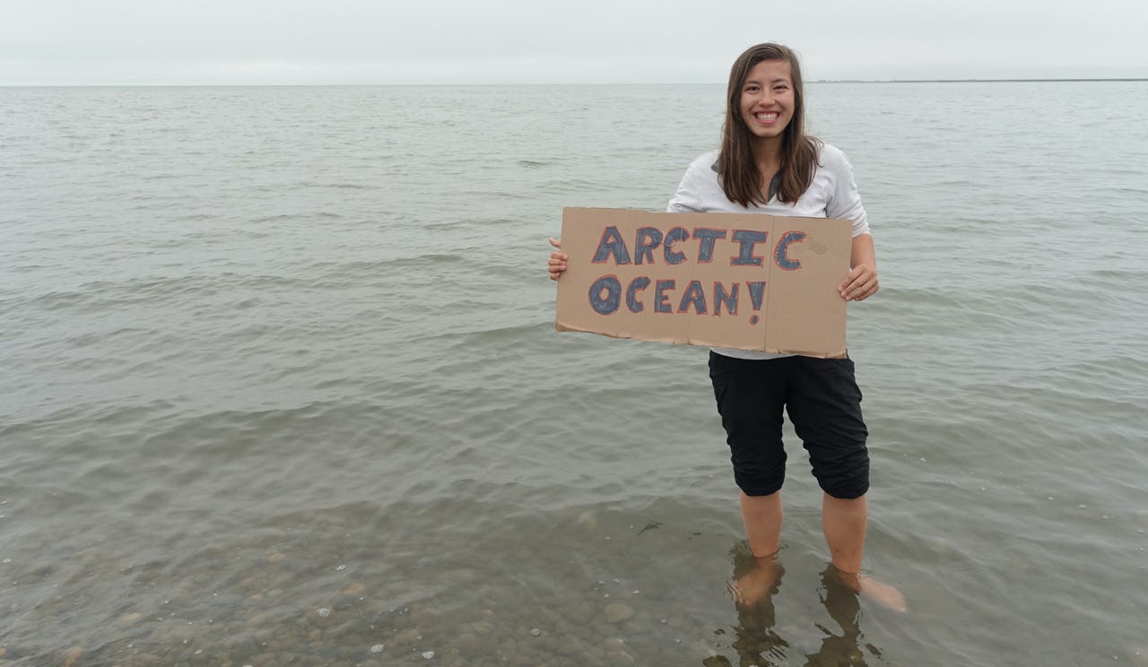Smiling woman with shoulder-length brown hair standing ankle-deep in the ocean with black trousers rolled up just below the knees and a light white long-sleeve shirt rolled up to the elbows holding a cardboard sign with “ARCTIC OCEAN!” written on it with the Arctic ocean in the background