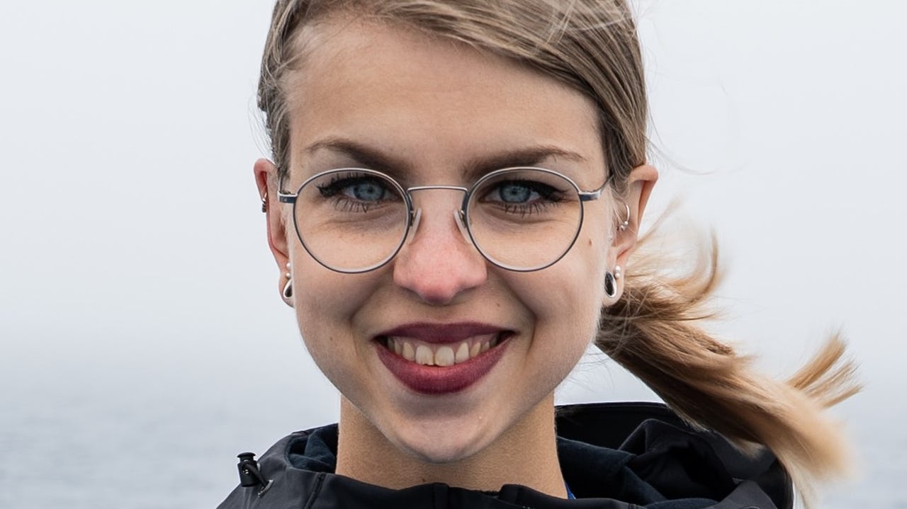 Close-up of a smiling woman with blond hair and round metal frame glasses with grey and blue sea in the background