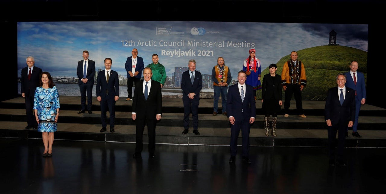 16 individuals wearing traditional suits and formal business attire standing 2 metres apart with a background display of the 12th Arctic Council Ministerial Meeting in Reykjavik, Iceland