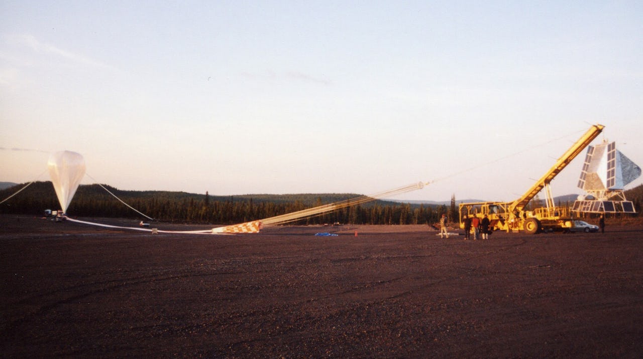 A white weather balloon attached to a yellow crane at a field with trees in the background