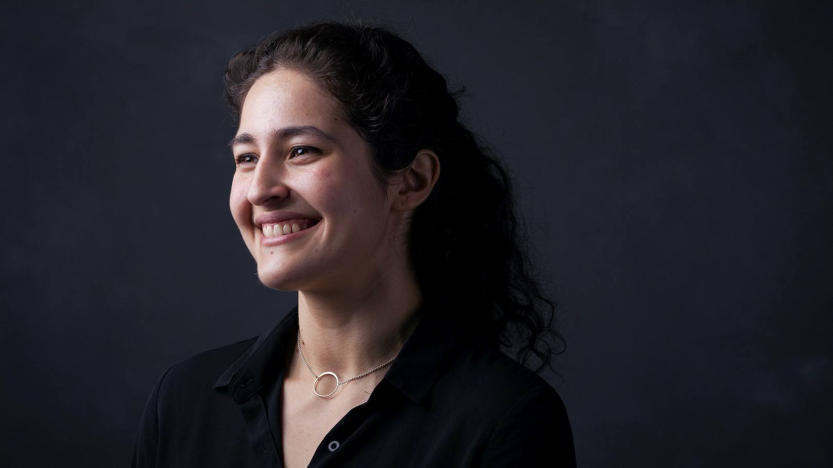 Portrait of a smiling woman with dark brown hair looking toward the left and wearing a black blouse and a golden ring necklace