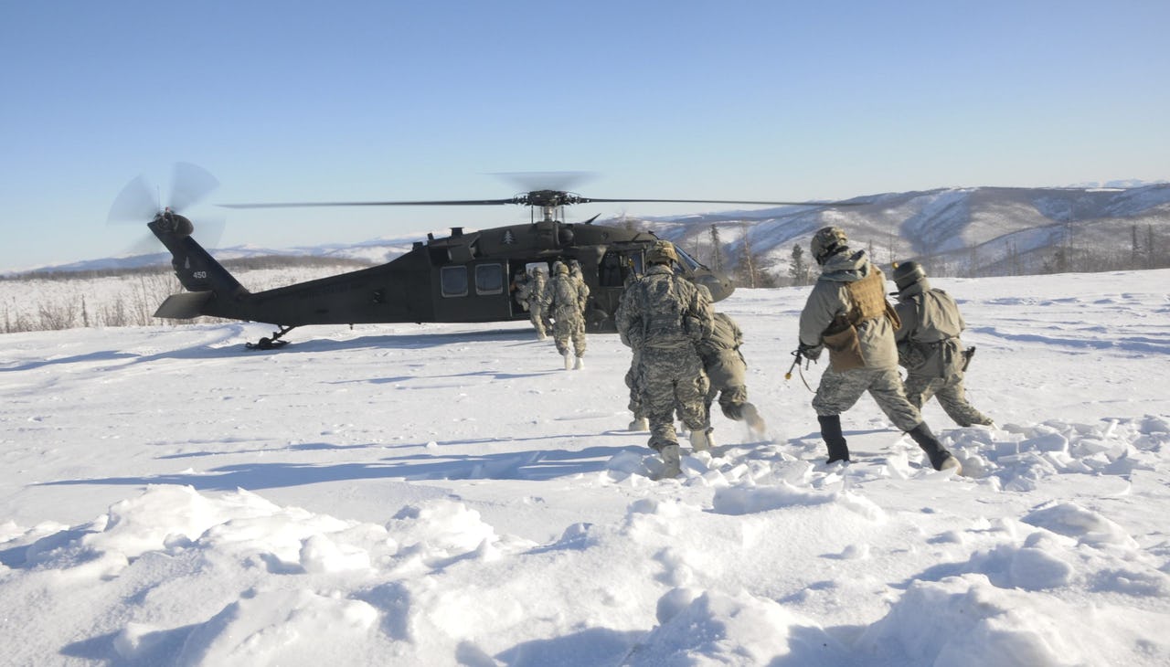 Military members dressed in grey fatigues run across snow towards a helicopter