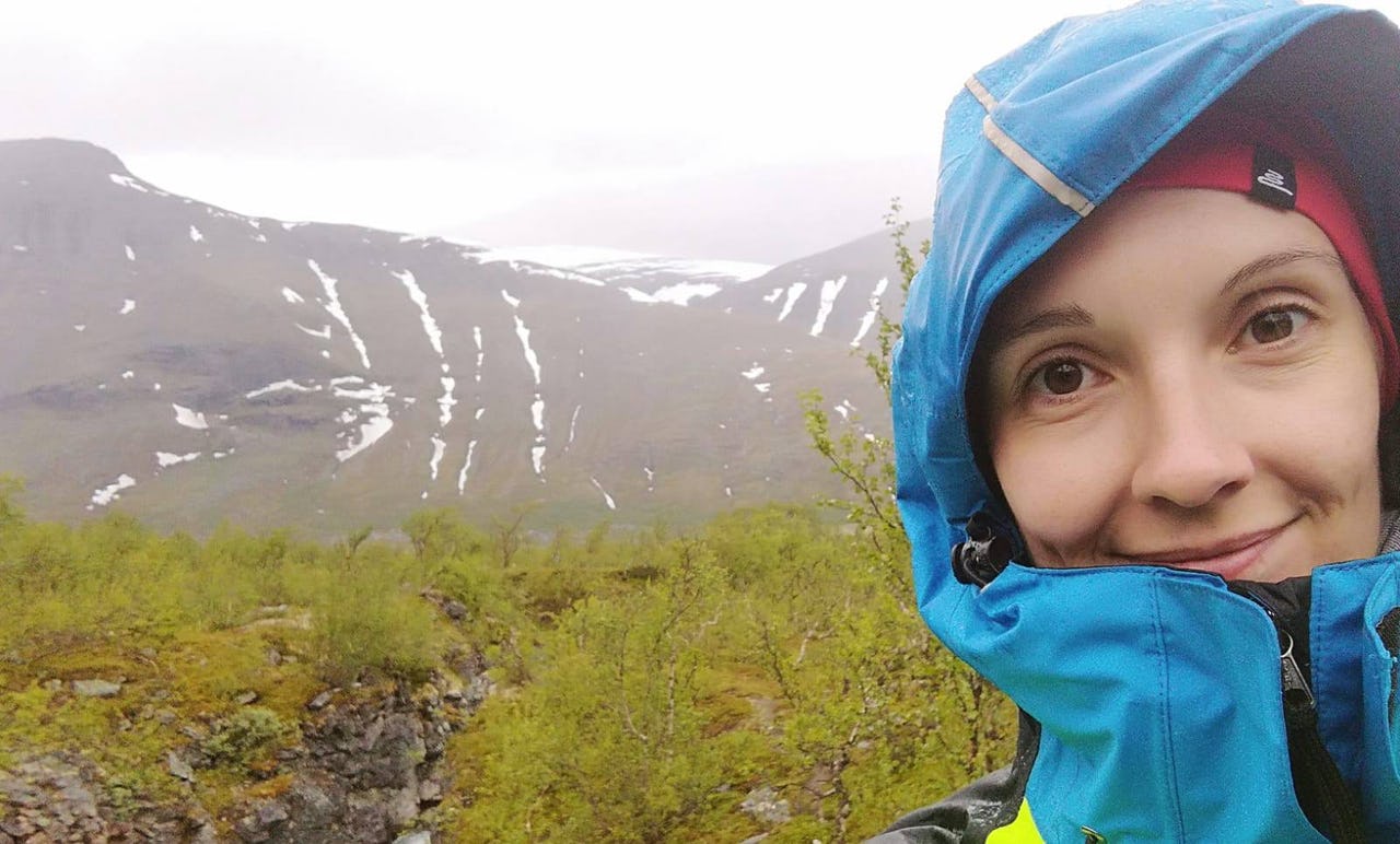 Smiling woman with blue and yellow hardshell rain jacket in an Arctic field with snowy mountains in the background