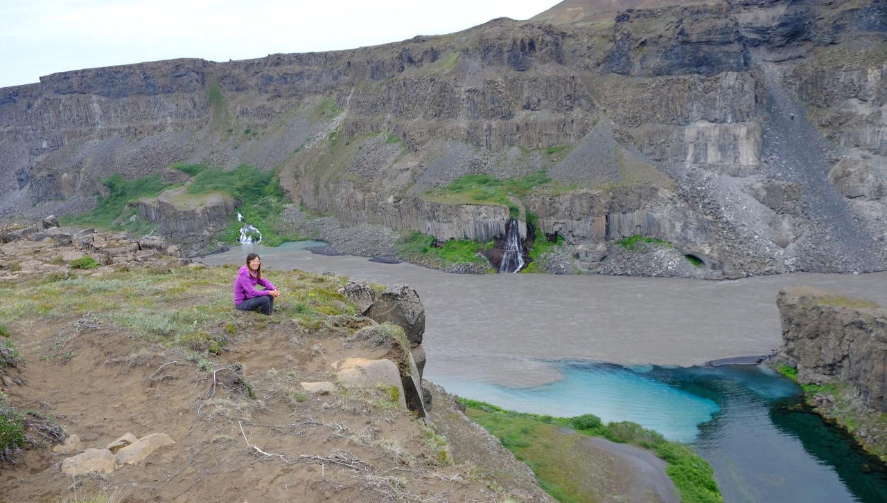Woman with purple technical jacket sat on mud and grass with Icelandic river and hills in the background