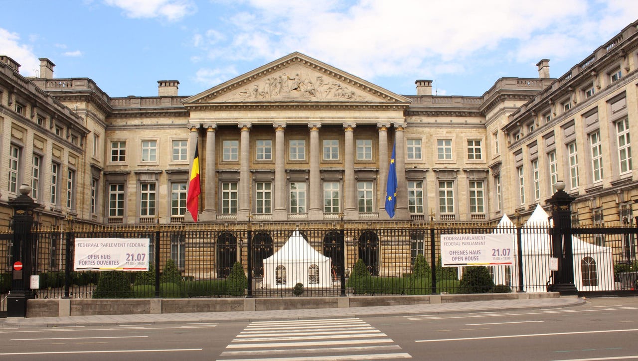 Neoclassical architecture parliamentary building with eight colonnades with black, yellow, and red (Belgium) flag on one side and a blue flag with yellow stars (European Union) on the other, and a garden. A black gate closes the area from the street in the foreground of the picture