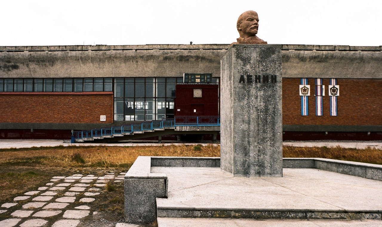 A dark grey bust of Lenin on a pedestal stands in front of a red brick building and grey-white sky