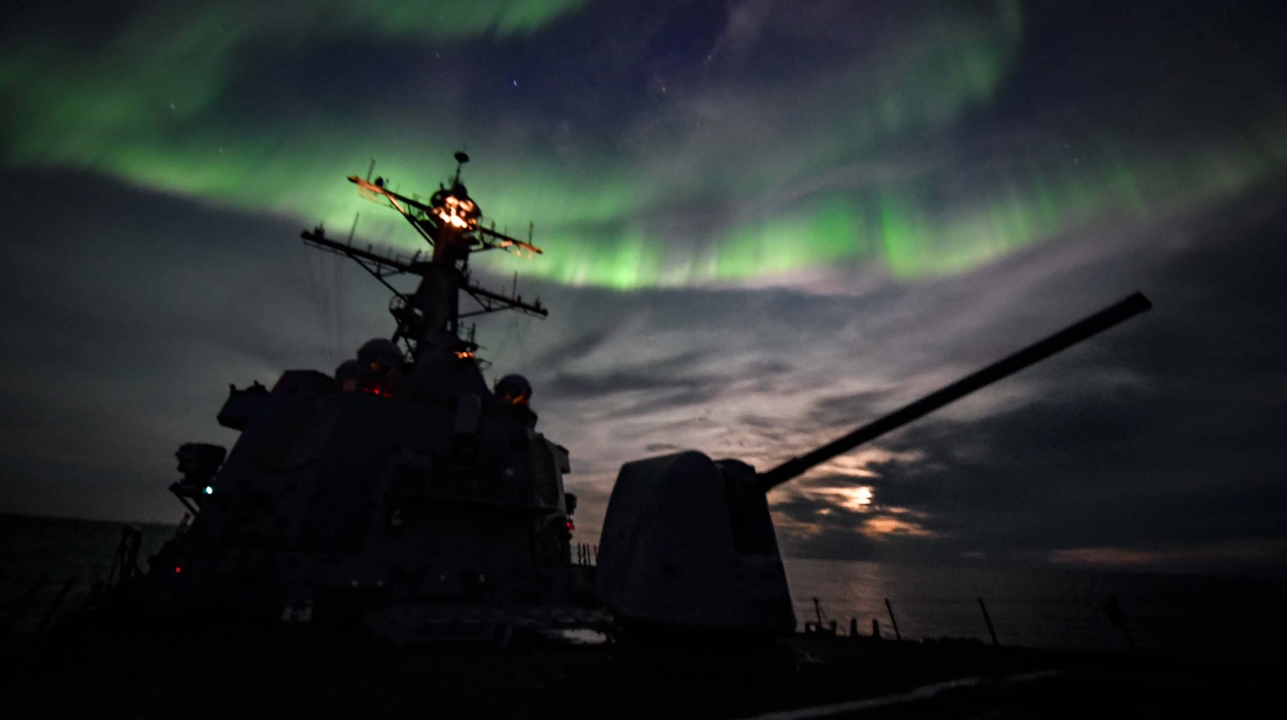A black silhouette of a military ship against a backdrop of green northern lights in the sky