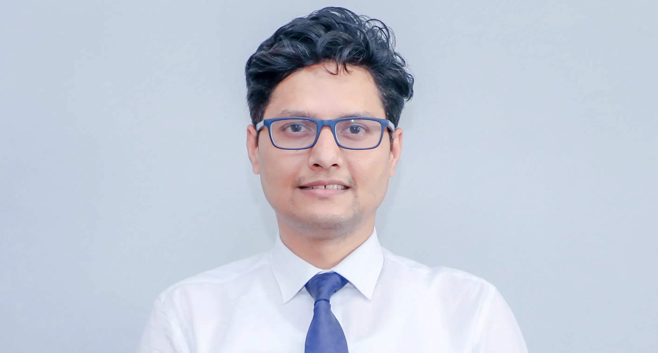 Man with blue glasses, shirt, and blue tie looking straight at the camera