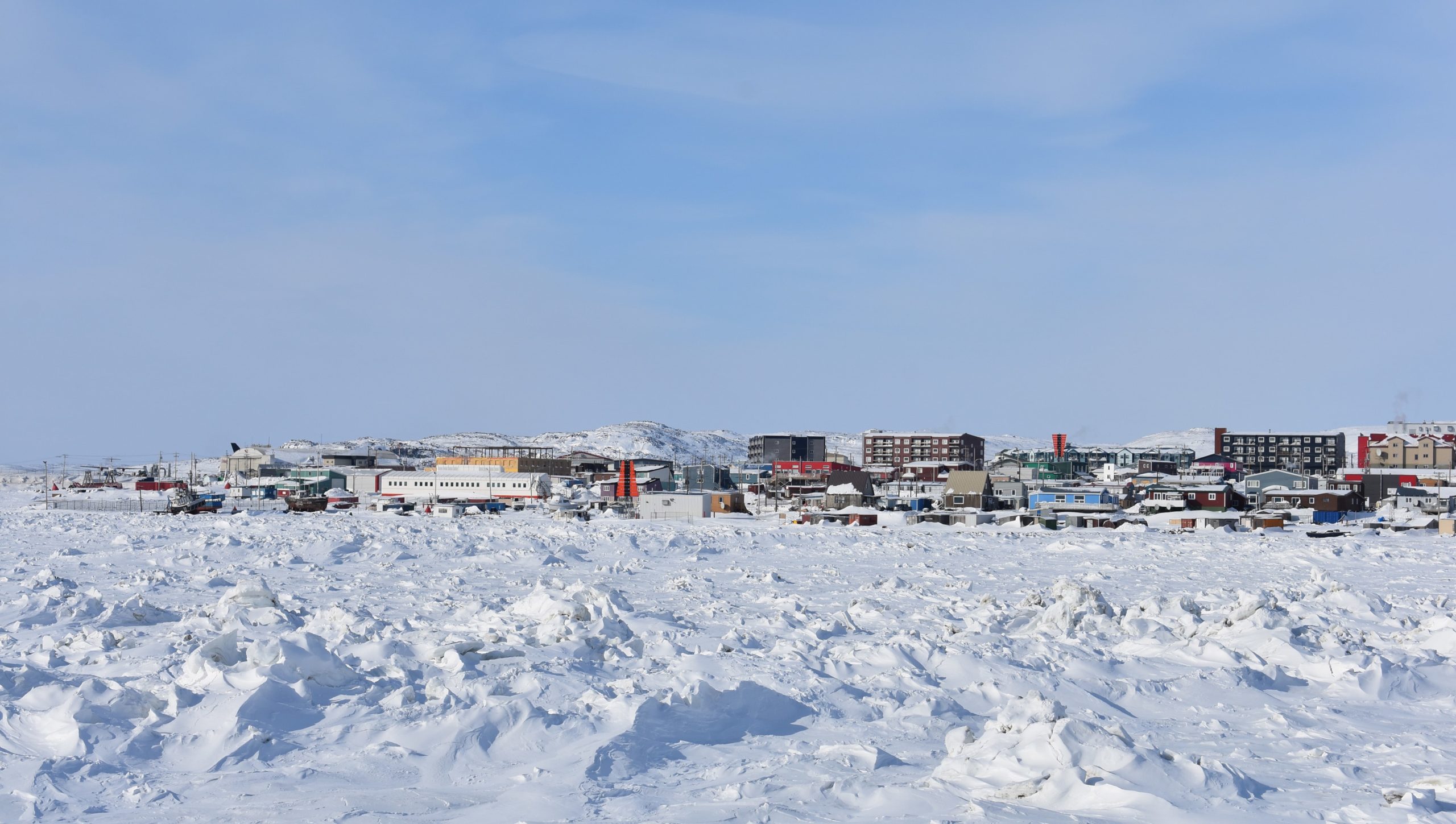 Image of Iqaluit from the Arctic Sea Ice in Spring 2021, colorful buildings can be seen along with a blue sky and snow-covered ice