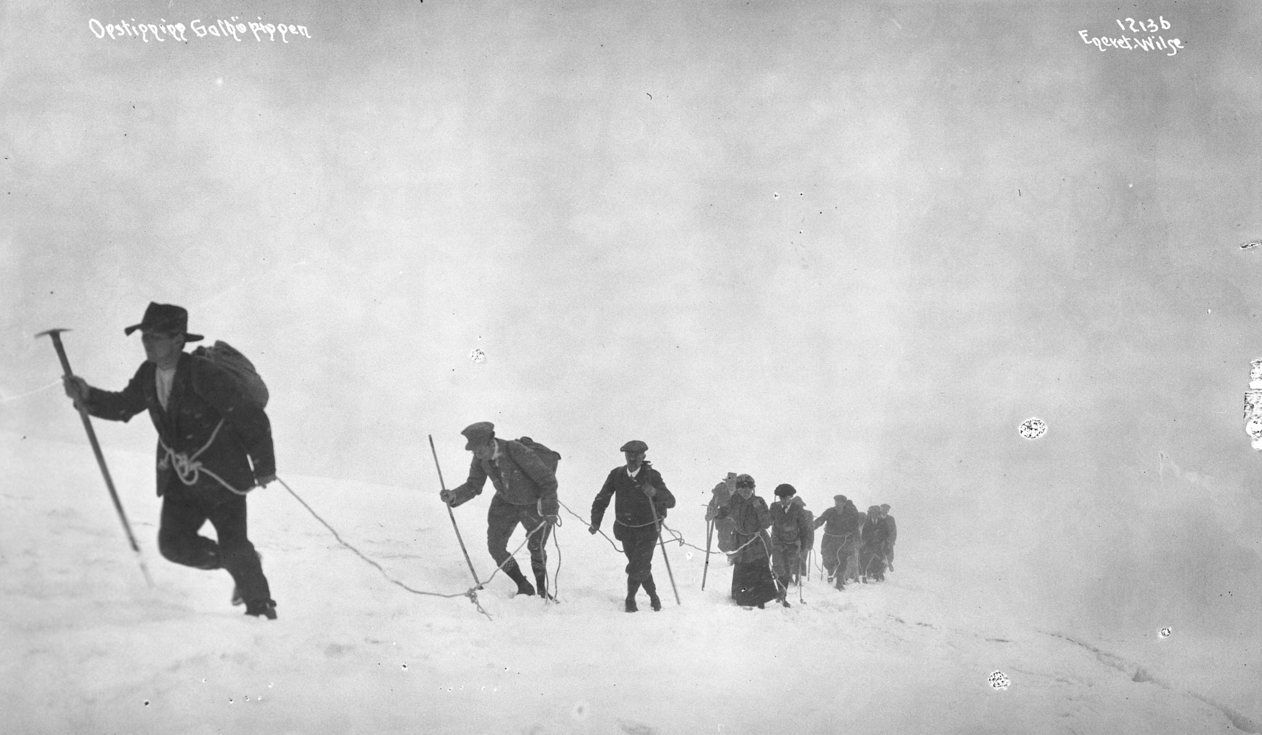 A black and white photograph of a group of around 10 mountaineers walking up a steep snowy mountainside