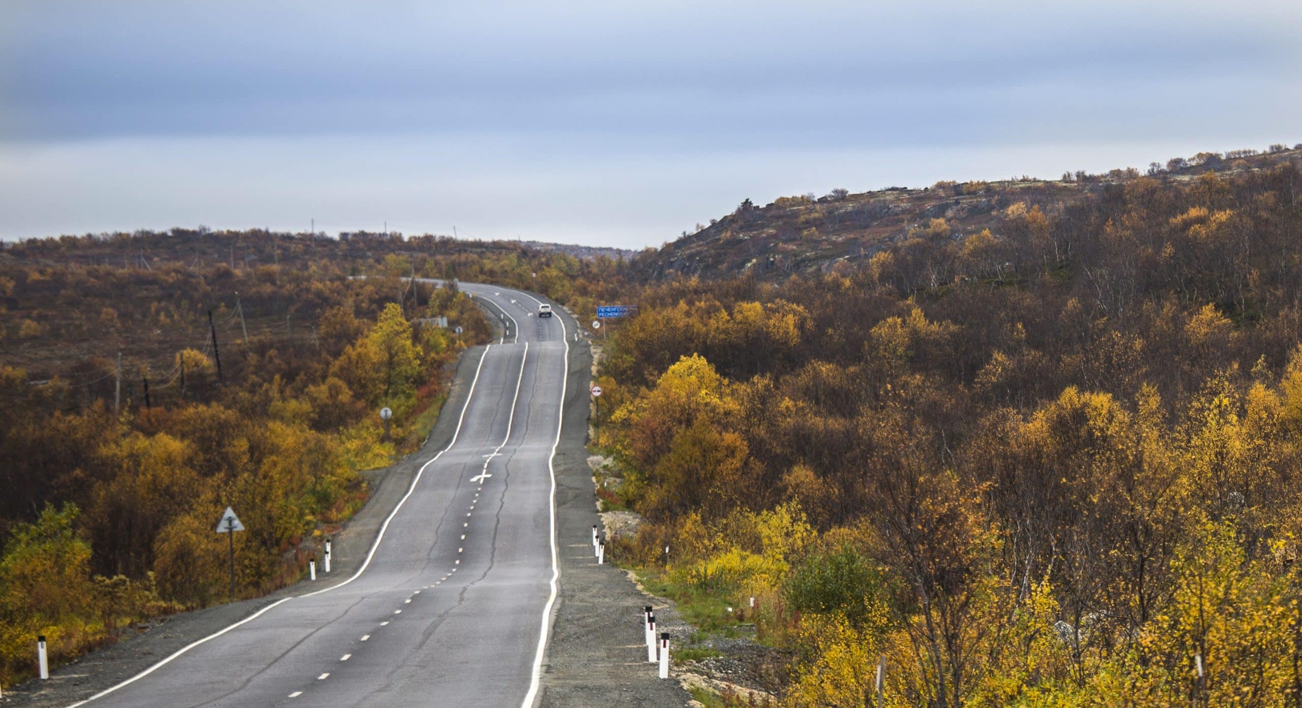 A car drives on a paved road on Kola Peninsula, Russia, in between low forests and shrubs in fall