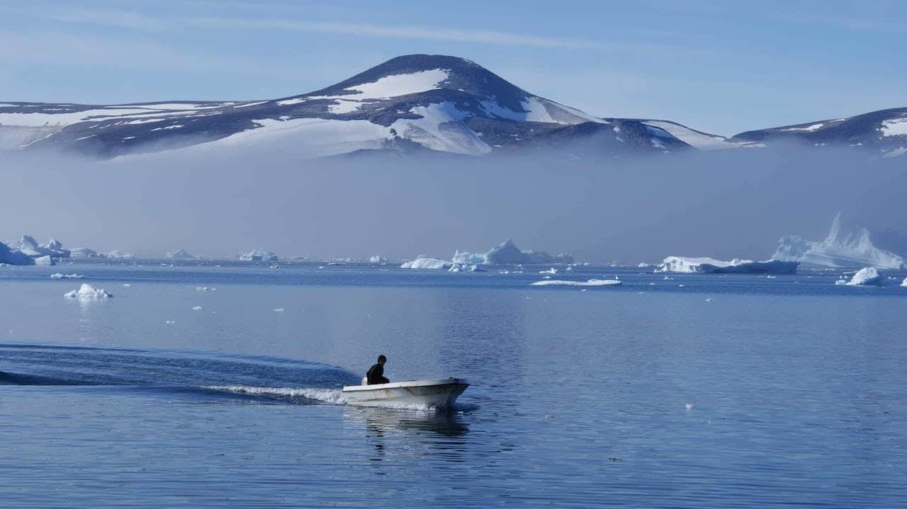 An Inuit hunter boating through Greenlandic waters with snow mountains and icebergs in the back