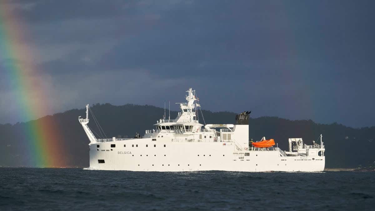 RV Belgica, a large white ship, at sea in gray water, sailing to the left towards a rainbow