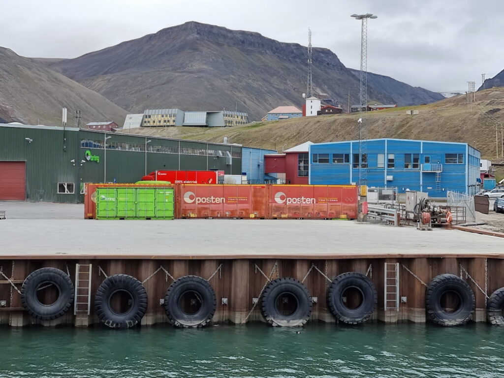 A waterfront industrial dock with large black tire bumpers lining the quay beside calm green water. In the foreground are orange shipping containers marked "Posten" and a bright green container. Behind them stand blue and dark green warehouse buildings, utility towers, and a few small houses on a hillside. Steep, rocky mountains rise in the background under an overcast sky, creating a stark Arctic landscape