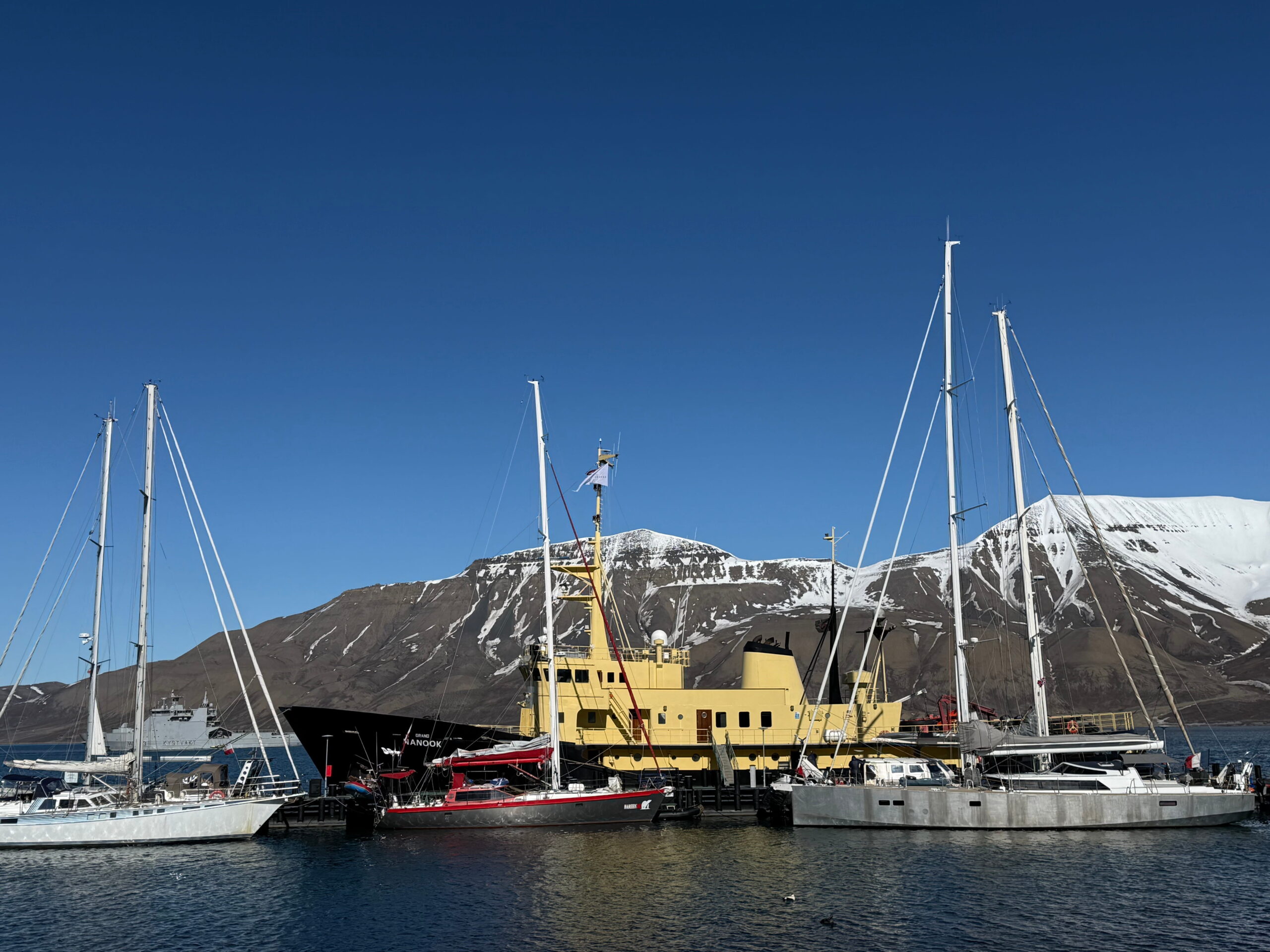 Sailboats and an expedition-style ship are docked in a harbor against a backdrop of snow-covered mountains under a clear blue sky