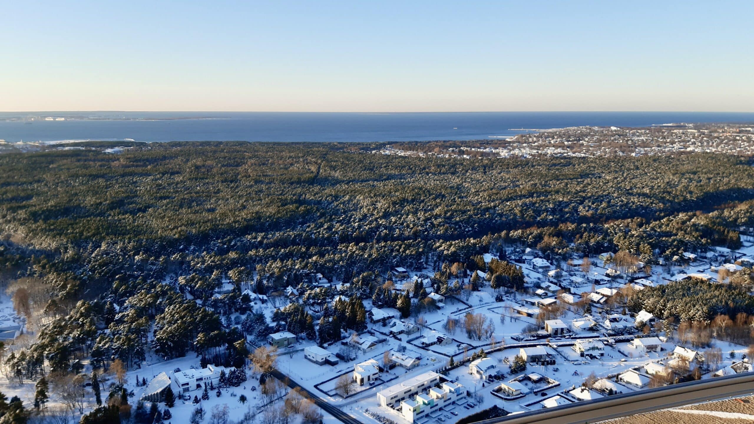 The photo depicts a sunny, clear-skied birds-eye view of the landscape with snowy residential buildings at the bottom of the image and, further in the distance, spruce forest and blue-colored water of Tallinn Bay
