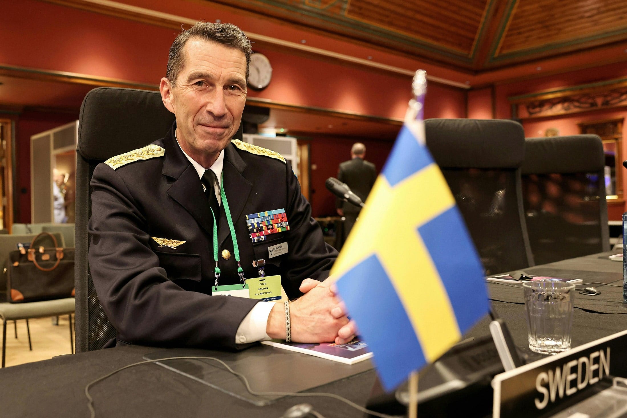 General Micael Bydén, Sweden’s Chief of Defence, with his hands on a brown table with a Swedish flag in front of him and a plaque that says “Sweden”