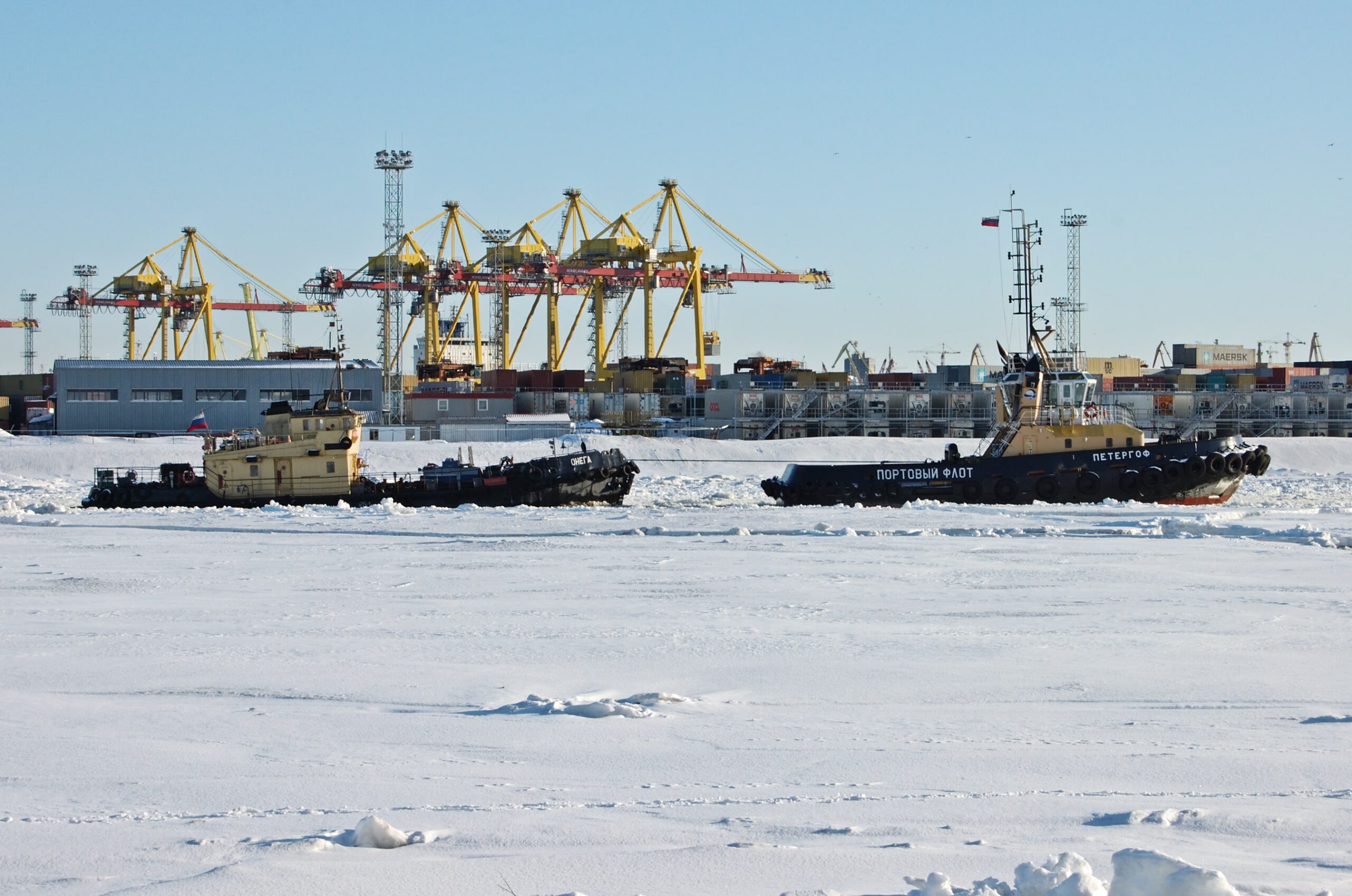 Two Russian boats in front of the container terminal within the Saint Petersburg seaport, surrounded by snow and ice