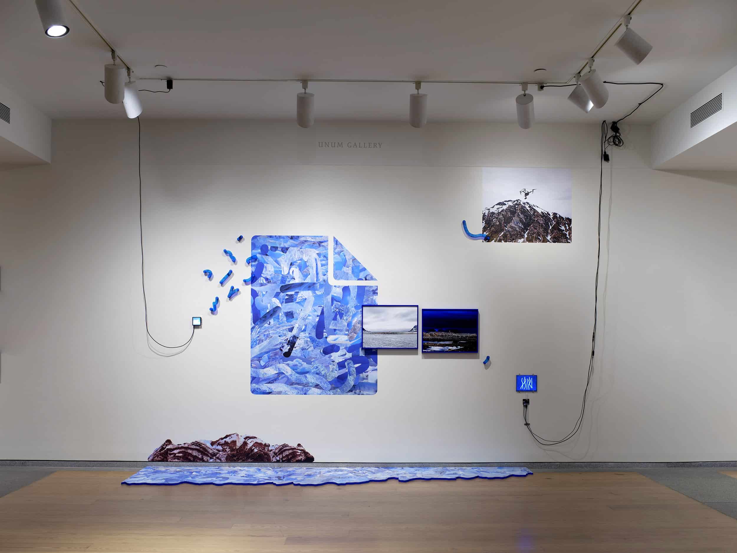 A multimedia installation showing combined images of ice in the shape of a folder, two small video pieces, two blue framed images of the Arctic landscape, an image of a drone flying in the sky, muddy ice, and a edge painted PVC floor piece that looks like a glacier.