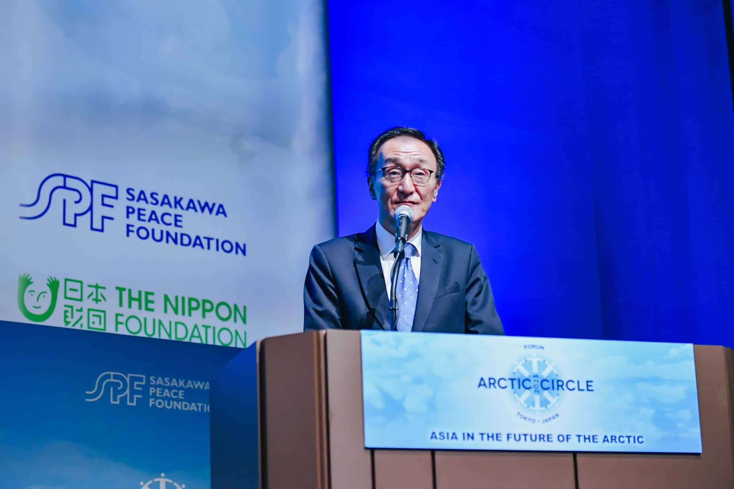 Keizo Takewaka speaks into a microphone at a podium with a dark blue and white background