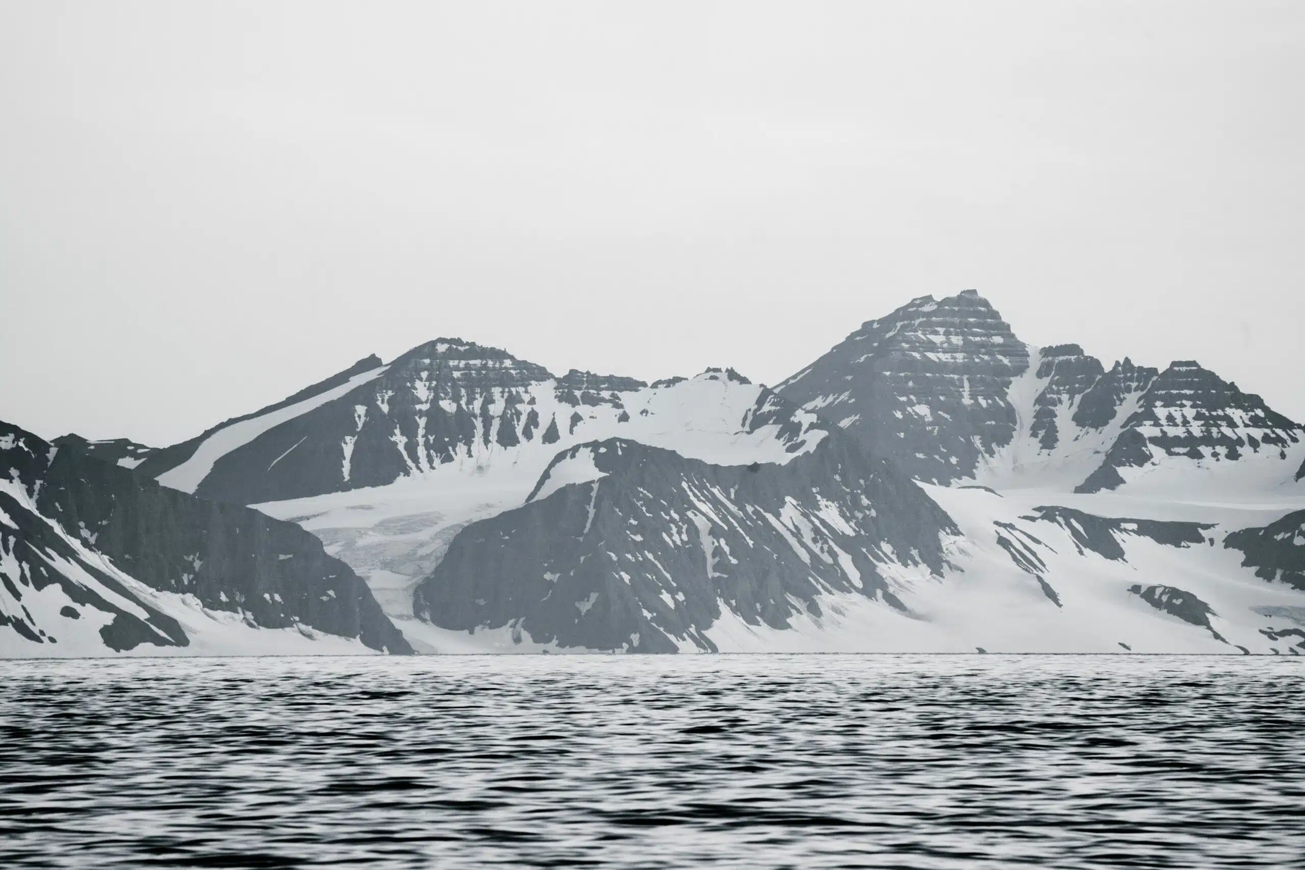 Gray snowy mountains against a pale gray sky with dark water in the foreground