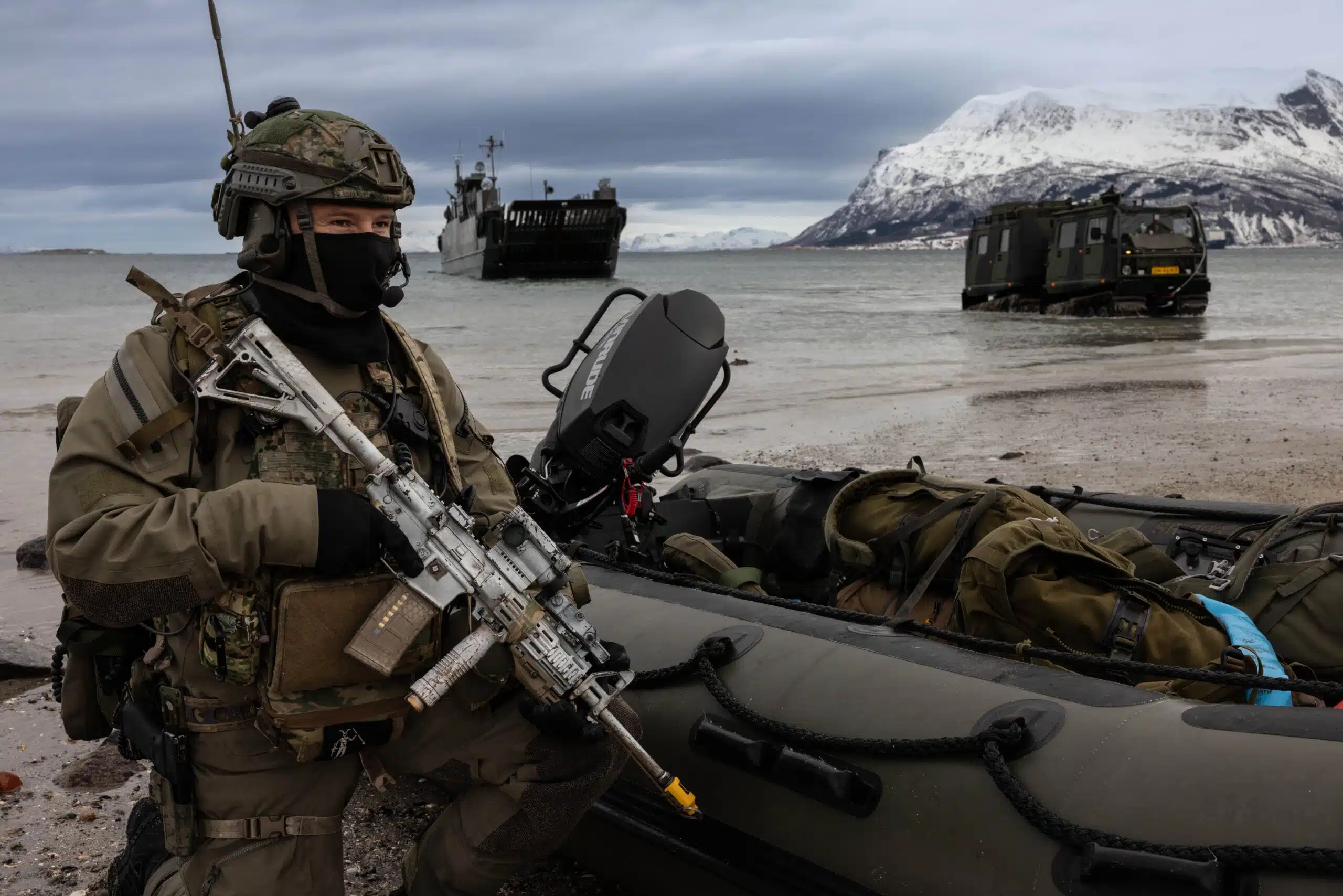 A soldier in a dark uniform with a grey rifle stands in front of brown water with brown military equipment and a grey sky in the background